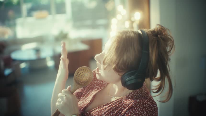 A young woman wearing large headphones sings loudly and dances while using a wooden spoon as a microphone in a kitchen