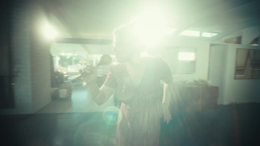 A young woman wearing large headphones sings loudly and dances while using a wooden spoon as a microphone in a kitchen
