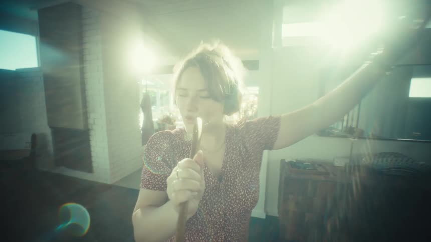 A young woman wearing large headphones sings loudly and dances while using a wooden spoon as a microphone in a kitchen
