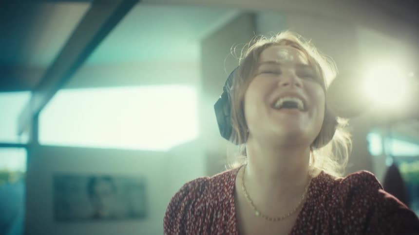 A young woman wearing large headphones sings loudly and dances while using a wooden spoon as a microphone in a kitchen