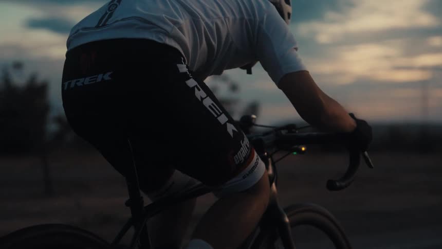 A view from behind shows a cyclist pedaling a road bike along a flat path during a dark sunset