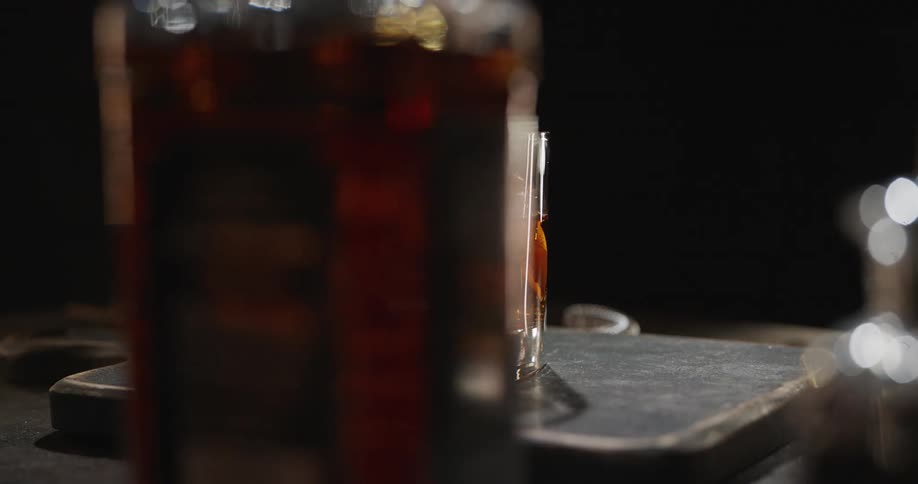 An old fashioned cocktail with a large clear ice cube orange twist and cherry sits in a rocks glass on a dark bar counter surrounded by a blurry shaker and strainer