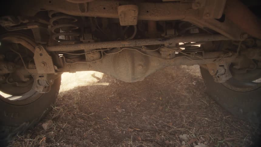 Underside of a vehicle shows axle suspension and wheels above dirt ground