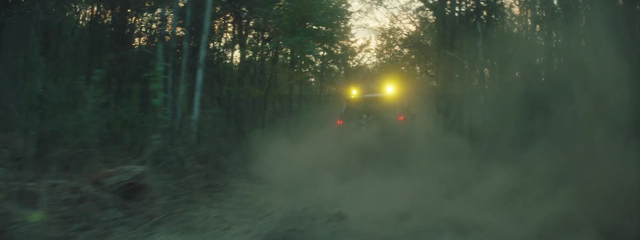 A tan vehicle with bright lights and mud on the exterior speeds through a dark forest and leaves a thick trail of dust behind