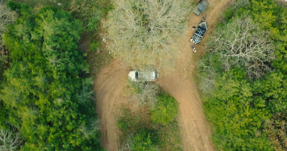 An aerial view shows a tan vehicle driving along a winding dirt path through a forest of green trees and bare branches