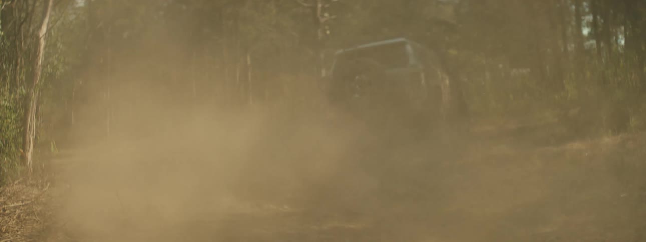 A tan vehicle drives quickly across dry grass and through a forest while creating a thick cloud of dust