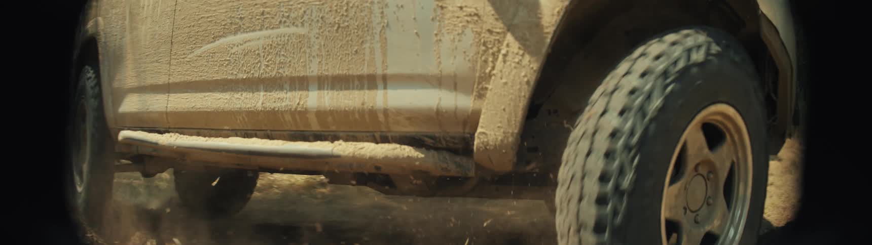 A muddy offroad vehicle drives quickly across a dirt path kicking up a large cloud of dust and debris the camera follows the side of the truck showing the mudsplattered wheels and body as it accelerates