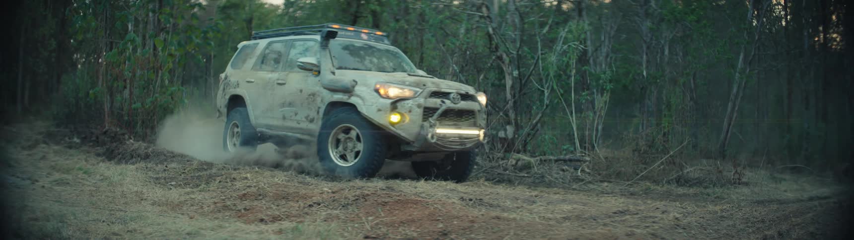 Truck with headlights on travels on a rural dirt road though the woods leaving behind a trail of smoke and dust