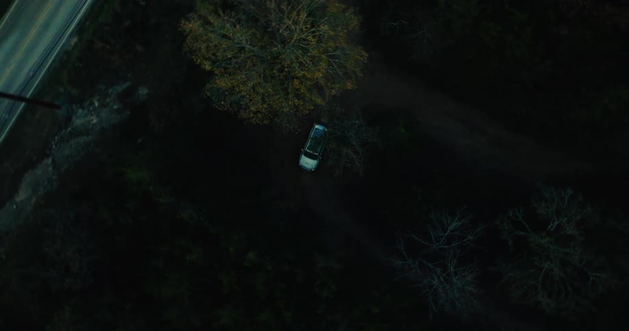 An aerial drone shot follows a lightcolored vehicle as it navigates a winding dirt path through a dark densely wooded areathe overhead perspective captures the vehicle moving slowly beneath the canopy of bare and evergreen trees during twilight