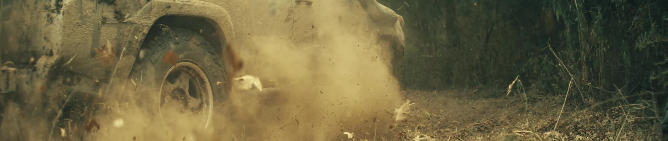A close ground level view shows a large tire spinning and kicking up dirt and dry straw as the vehicle drives forward