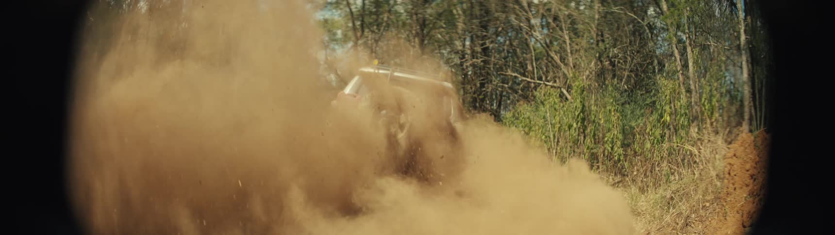 A low angle shot shows the wheels of a tan vehicle as they spin rapidly and throw dirt and thick dust into the air while the car speeds away through a wooded area