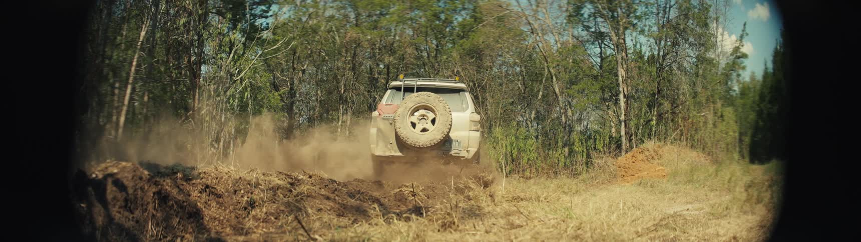 A tan mud splattered vehicle with a roof rack and snorkel drives quickly across a field of dry grass and dirt while kicking up a thick cloud of dust