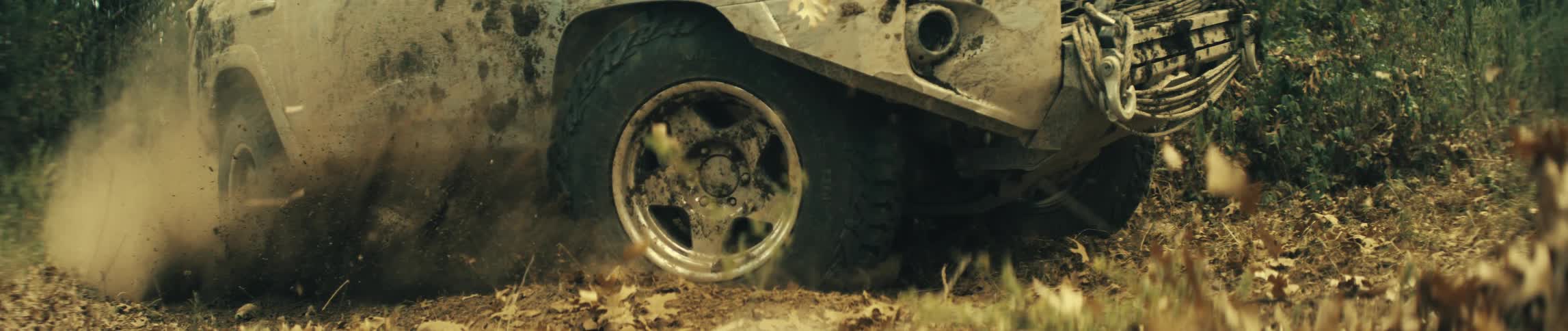 A close ground level view shows the front wheels of a mud covered vehicle spinning and kicking up dirt and dry leaves as it drives forward