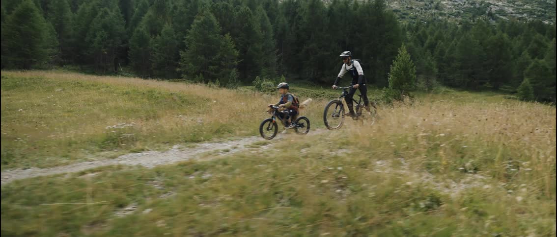 A child and his father riding bikes on a trail in the woods as a dog runs along with them at midday