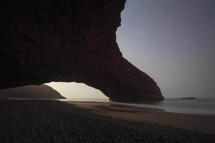A time lapse shot captures a star filled sky over a rocky beach archway at dusk