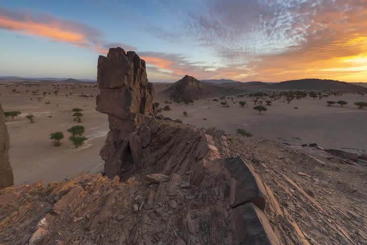 A tall stone pillar stands prominently on a rocky ridge as the morning sun rises over a vast desert plain dotted with small trees and distant mountains under a colorful sky filled with textured clouds