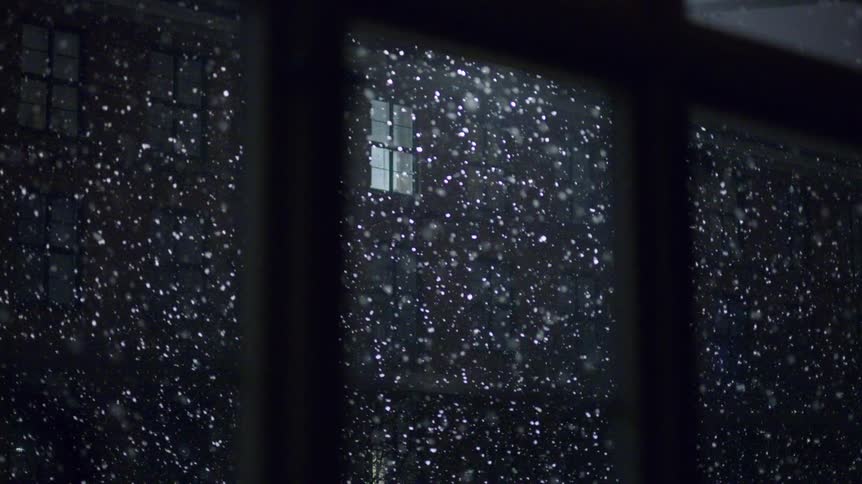 Snow falling in front of a window with view of a lit building at night