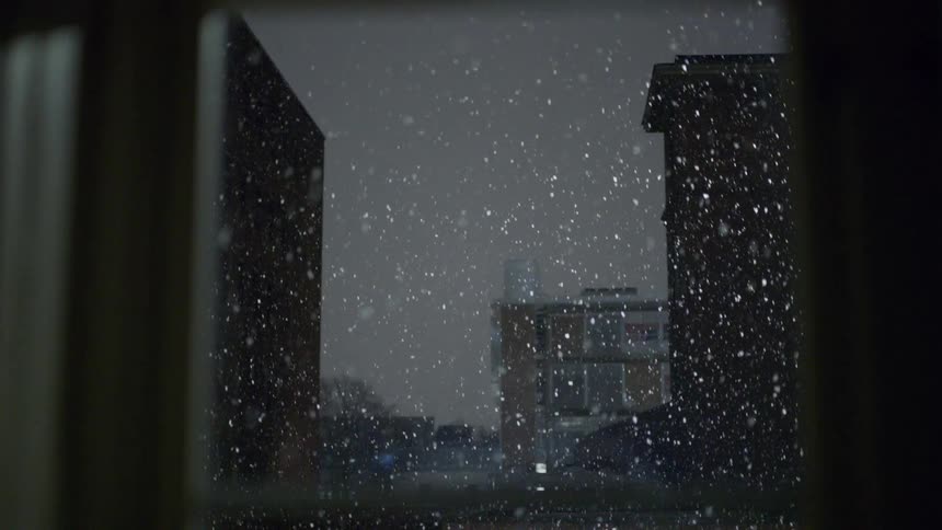 Heavy snow falling at night above a dark city skyline with glowing city buildings