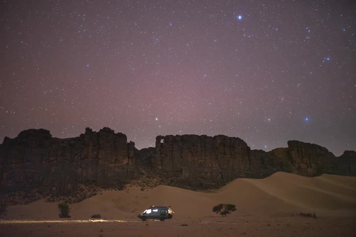 A lone van with headlights on is parked in a vast desert under a starry night sky