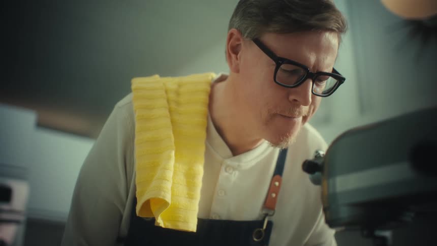 A baker wearing glasses and an apron operating a silver stand mixer in a modern kitchen