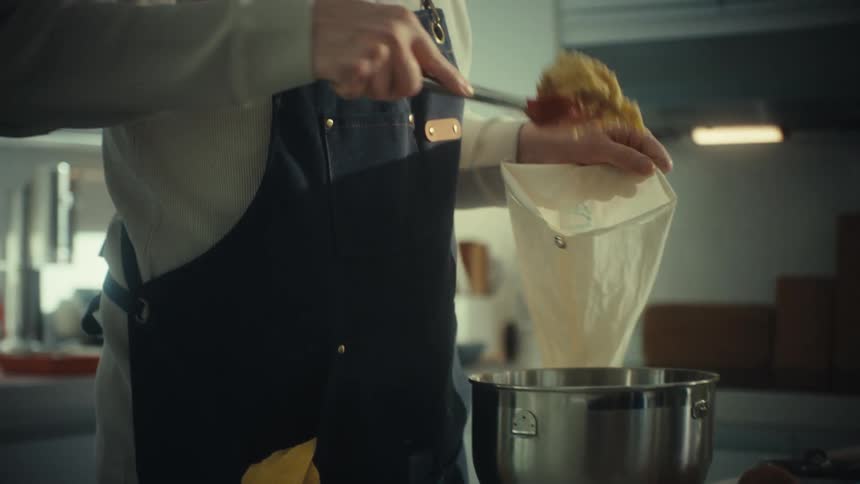 A person wearing an apron pours ingredients from a cloth bag into a pot in a kitchen