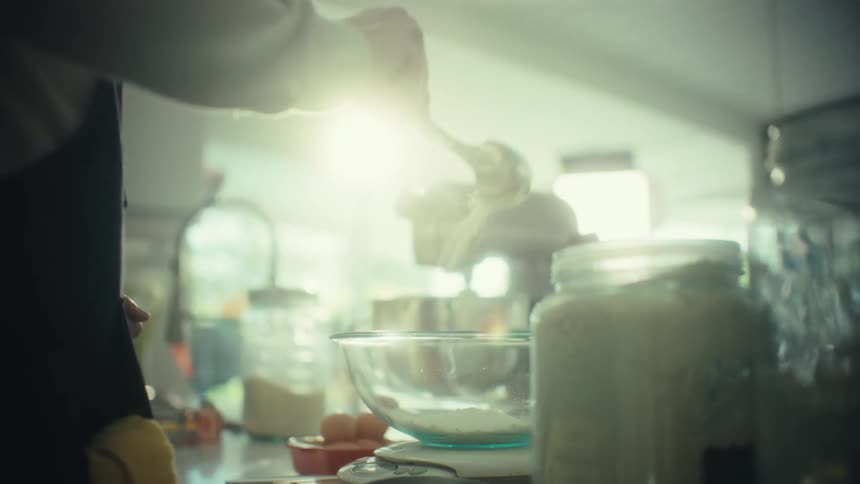 A person wearing a dark apron pouring flour from a container into a glass bowl in a bright sunlit kitchen