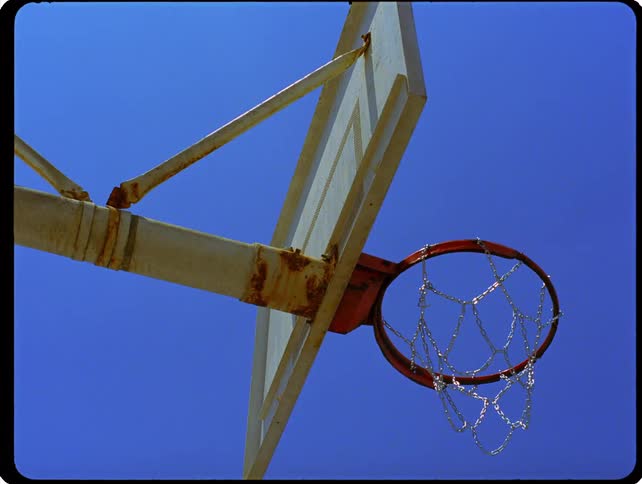 A low angle static shot shows a basketball hoop with a rusty rim and a chain net under a clear blue sky