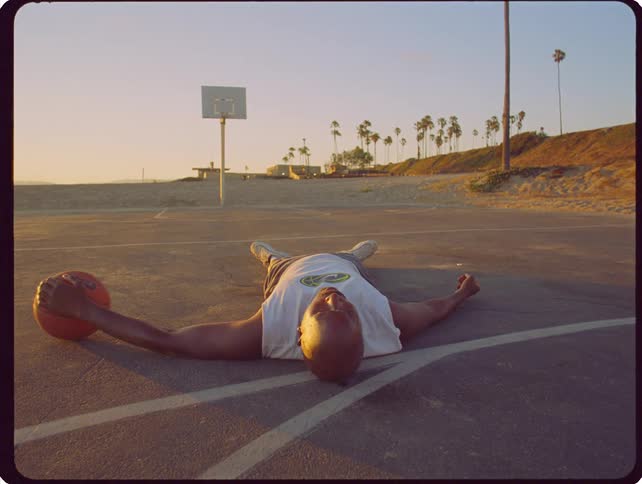 A man lies flat on his back in the middle of an outdoor basketball court while holding a basketball during a golden sunset at the beach