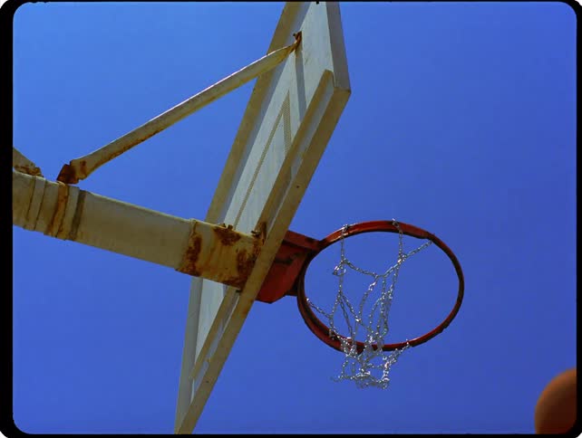A view from below shows a basketball bouncing off the rim of an outdoor hoop with a chain net under a clear blue sky