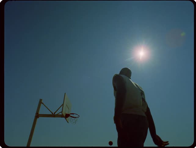 A person stands on a basketball court and shoots up at a basketball hoop under a bright sun in a clear blue sky