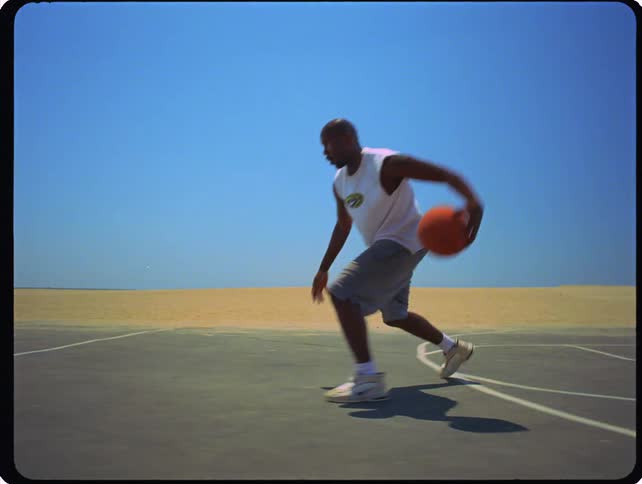 A man wearing a white tank top and grey shorts dribbles a basketball across an outdoor court located next to a sandy beach under a clear blue sky