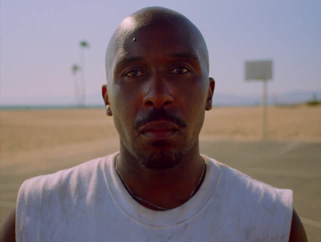 A man stares directly into the lens with a stoic expression while standing on an outdoor basketball court the sun casting soft light over his features with a beach and palm trees in the background
