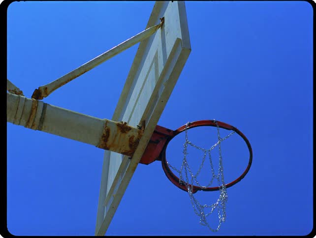 A low angle static shot shows a basketball hoop with a rusty rim and a chain net under a clear blue sky
