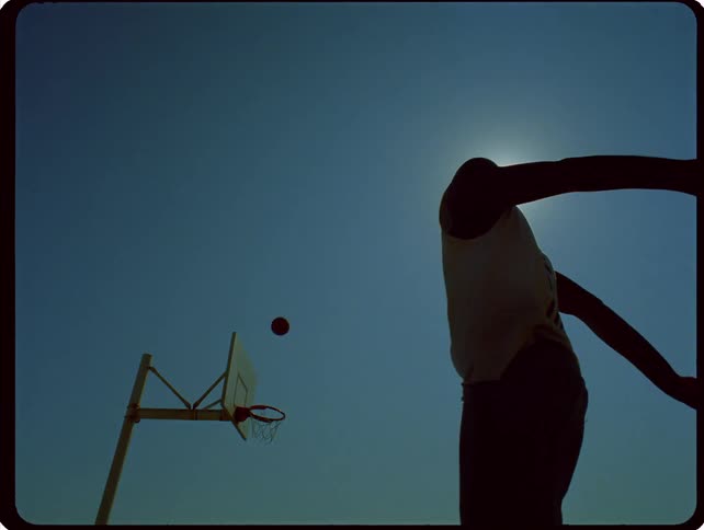 Silhouette of a person playing basketball on an outdoor court against a bright sunlit sky