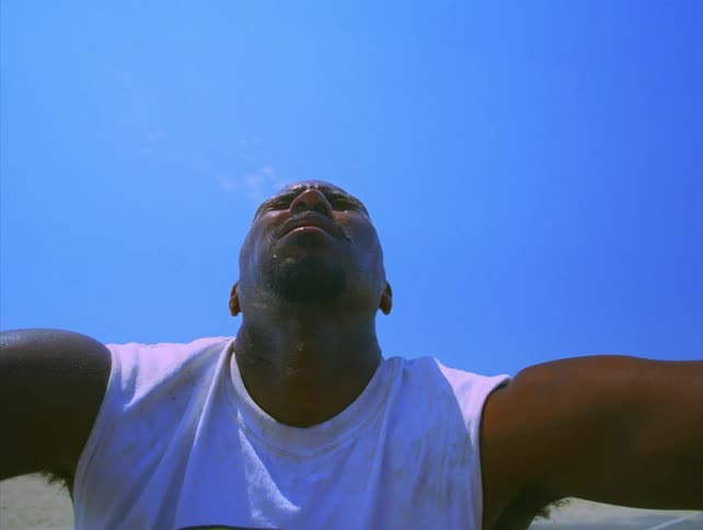 Man wearing a sleeveless shirt covers his face with his hand under a blue sky