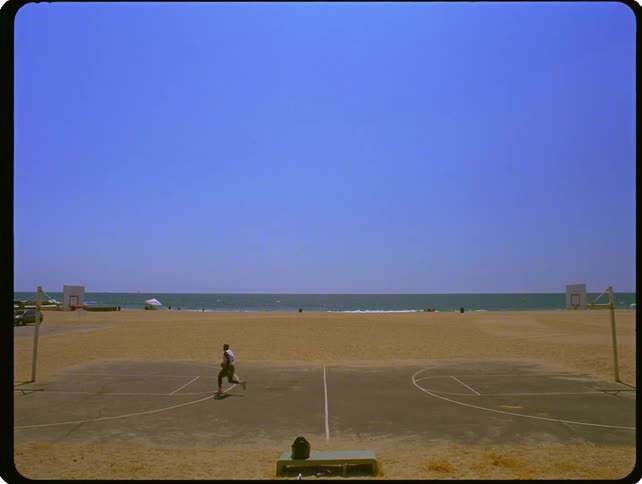 A wide shot shows a person dribbling a basketball across a full court located on a beach next to the ocean under a clear blue sky