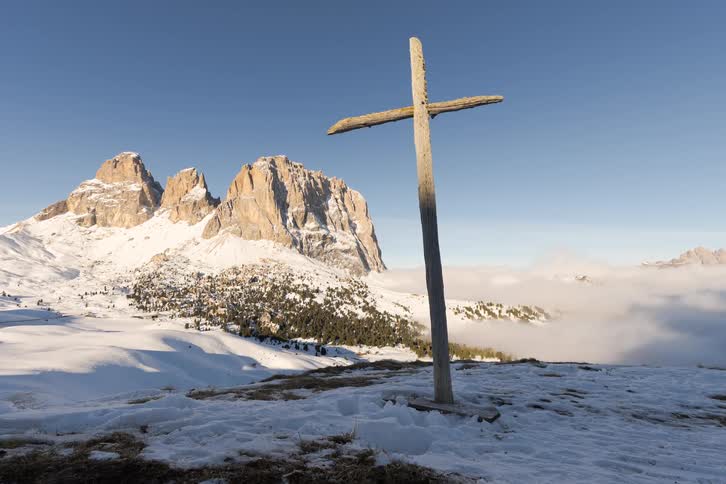 A large wooden cross stands prominently on a snowcovered mountain ridge as clouds drift through the valley below and sunlight illuminates the jagged rocky peaks in the distance