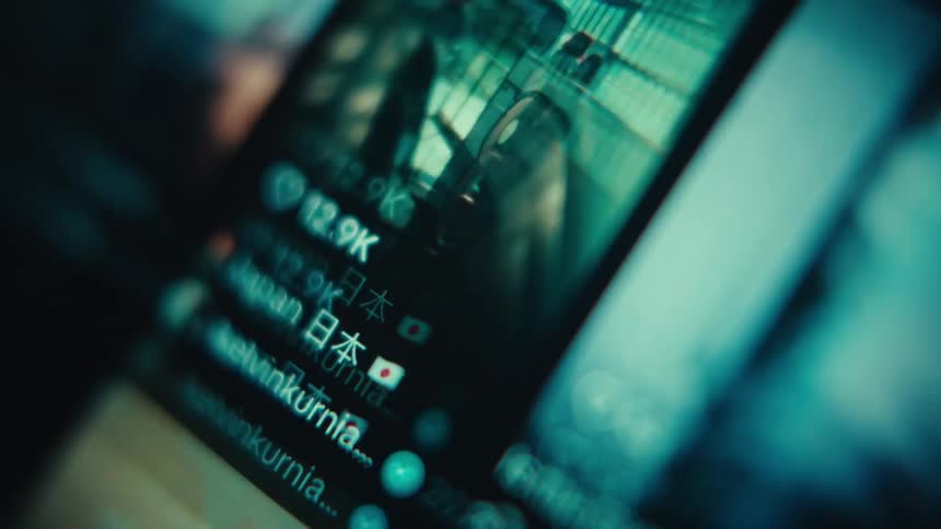 An extreme close up shot at a tilted angle shows a digital screen with a shallow depth of field as various social media video thumbnails and user names scroll quickly past the camera