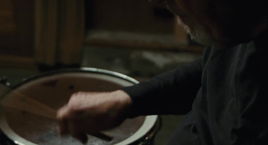 An elderly man with glasses and headphones plays a snare drum using brushes the camera moves from a closeup of his hands to a side profile of his focused expression