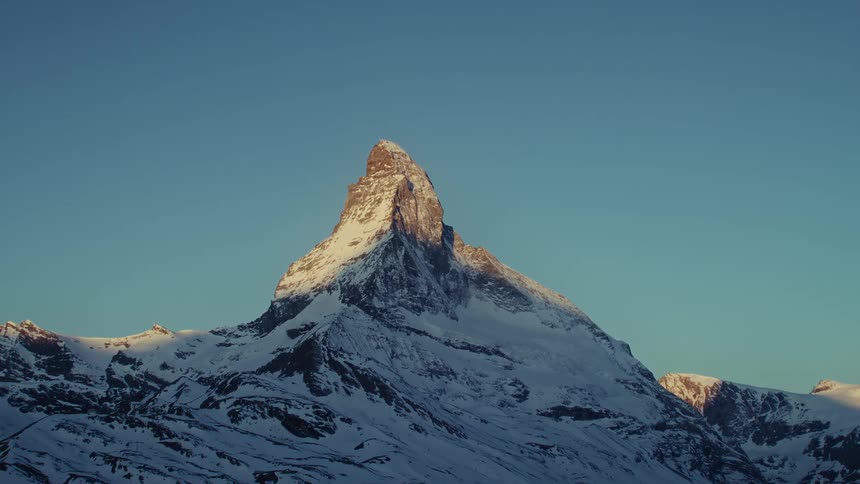 The sharp peak of a tall mountain covered in snow catches the warm golden light of a sunrise against a clear blue sky in a cold alpine environment