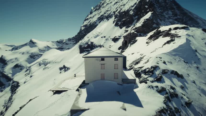 An Aerial Drone Shot of the Hörnli Hut Perched on a Narrow Snowy Ridge Surrounded by Jagged Mountain Peaks Under a Clear Blue Sky