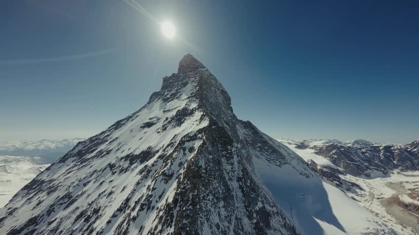 Snow on the rough terrain of a mountain peak with mountains in the distance as the sun shines brightly overhead
