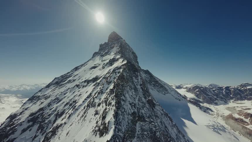 The Matterhorn covered in snow and ice under a bright sun and clear blue sky with surrounding mountain ranges and deep glacial valleys stretching toward the horizon