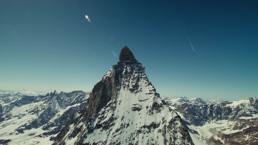 A drone push in to the Matterhorn rising above a vast snow covered alpine range under a clear blue sky as bright sunlight creates a lens flare across the frame
