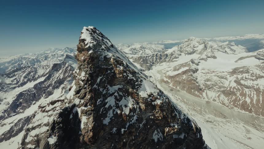An fpv drone flies over a sharp mountain ridge and moves toward a jagged peak in a snow covered range