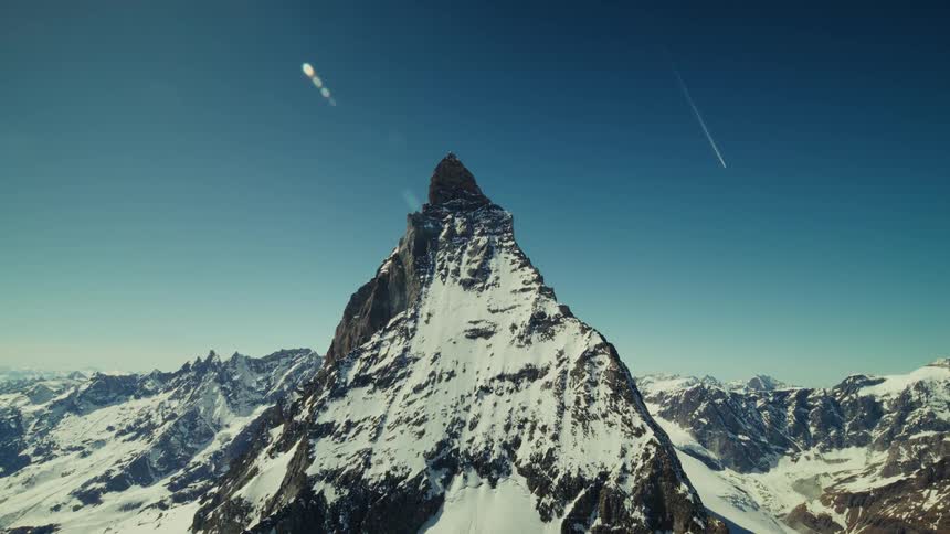 Mountain with a pointed peak and snow on the slope as the sun shines brightly in the sky as a jet airplane flies in the distance