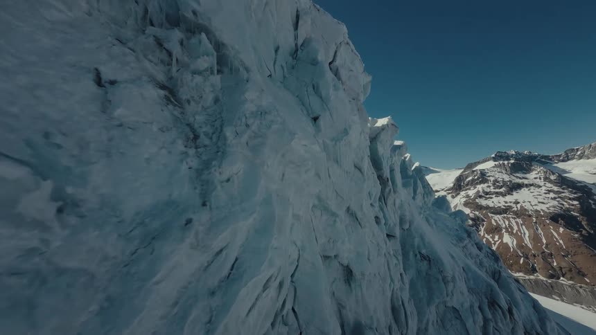 A drone flies closely alongside a massive jagged blue ice glacier revealing a steep mountain range with rocky brown peaks under a clear sky