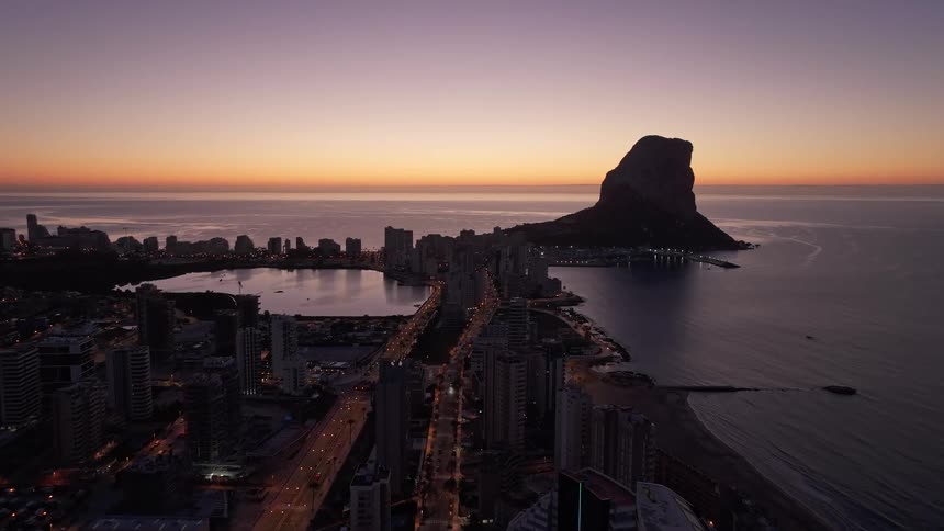 Illuminated avenue crossing a coastal urban area with a large rock formation in the sea at sunset