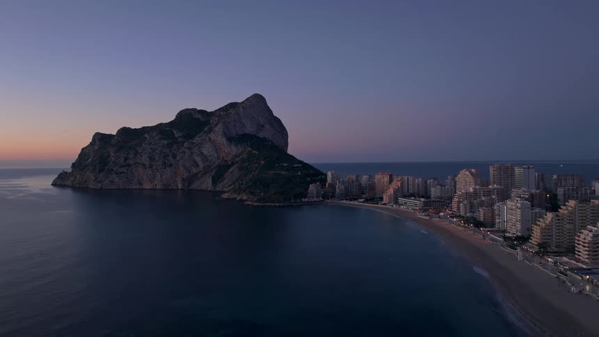An aerial view shows a large limestone rock formation rising from the sea next to a coastal city with tall buildings and a sandy beach under a dark purple sky at dusk