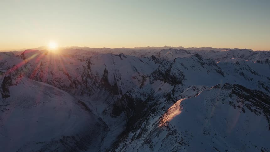 A wide aerial view shows the sun rising over a vast range of snow covered peaks in the alps during a clear morning with bright light and orange glows hitting the rugged winter landscape
